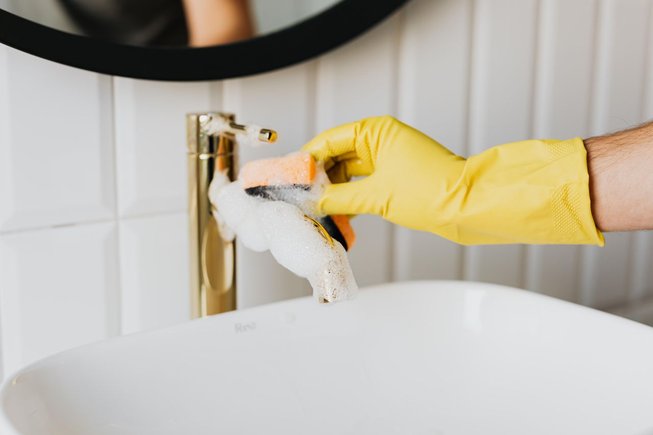 An unidentifiable man wearing yellow latex gloves is cleaning a gold faucet in a bathroom, using an orange sponge and detergent while tidying the home